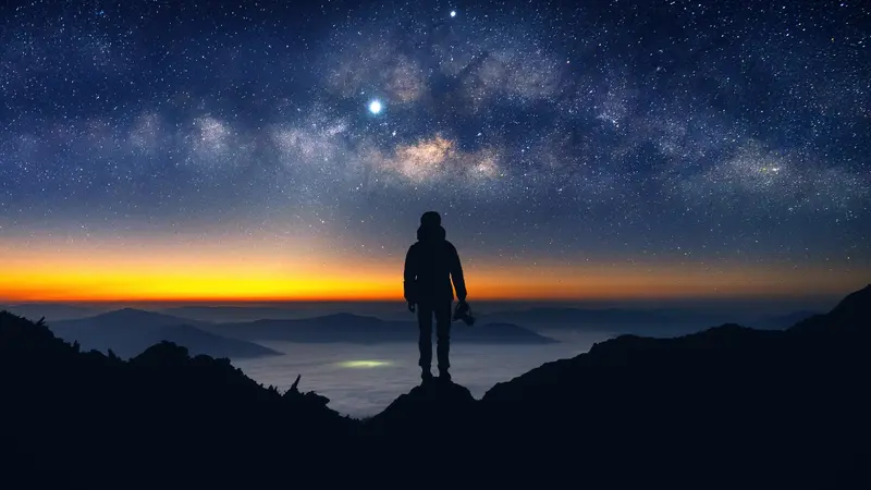 A silhouette of a person standing on a rock, at night, looking into the stars and sunset. 