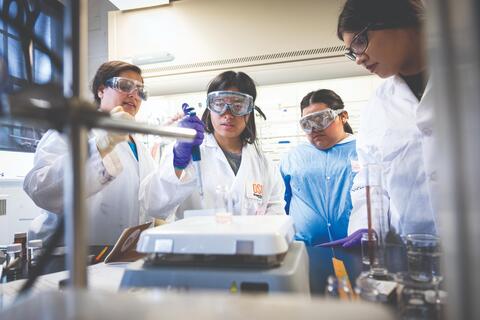 Students in lab coats and protective eyewear conduct an experiment in a science lab.