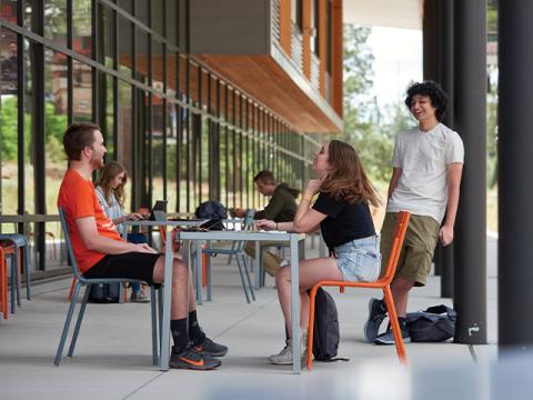 Two people sitting at table in foreground; third person leans against black pillar behind and to right.